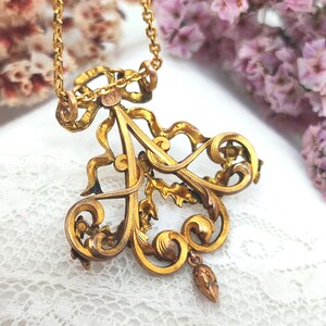 May include: A vintage gold-toned pendant necklace. The pendant features an ornate, stylized letter "A" with intricate scrollwork, a bow, and a small dangling element. The chain is a delicate link style. The background includes lace and flowers.