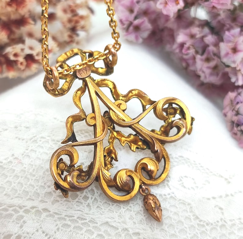 May include: A vintage gold-toned pendant necklace. The pendant features an ornate, stylized letter "A" with intricate scrollwork, a bow, and a small dangling element. The chain is a delicate link style. The background includes lace and flowers.