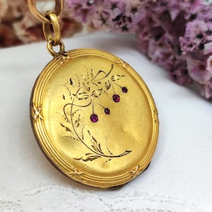 Op de afbeelding: Een goudkleurig medaillon met een bloemmotief en drie kleine, ronde, rode edelstenen. Het ronde medaillon heeft een decoratieve rand en hangt aan een gouden ketting. De achtergrond bevat paarse bloemen.
