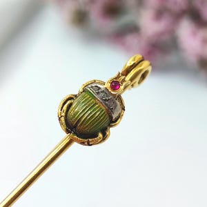 Op de afbeelding: Gouden haarspeld met een groen keverontwerp. De kever heeft een geribbelde textuur en is versierd met een zilveren band en kleine diamanten. Bovenop zit een kleine rode edelsteen.
