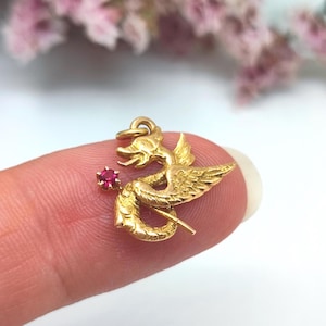 May include: A gold-toned metal charm shaped like a mythical dragon with textured wings and a coiled tail. The dragon figure features a small, round, faceted red gemstone set near its head and includes a circular loop at the top for attachment.
