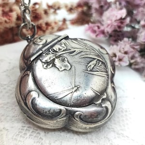 May include: A silver-toned metal locket with a rounded, scalloped edge design. The front surface features an embossed relief of a dragonfly hovering near tall grass and floral elements. A metal jump ring is attached to the top of the locket.