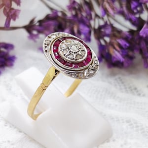 以下が含まれることがあります： 丸い中心の石が、小さな赤い石のハローで囲まれた、金のリング。リングは、小さな白い石の繊細なデザインでセットされています。