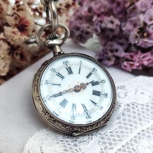 Puede incluir: Reloj de bolsillo plateado antiguo con esfera blanca, números romanos negros y agujas doradas. El reloj tiene un bisel grabado y una cadena. Flores secas y encaje en el fondo.