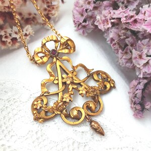 May include: A gold-toned pendant necklace featuring an ornate design. The pendant includes a bow, the letter "A", floral accents, and a small red gemstone. The necklace is displayed on a white surface with pink flowers in the background.