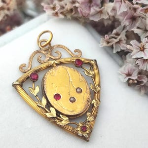 Può includere: Un ciondolo triangolare color oro con un elemento centrale ovale caratterizzato da linee incise. La cornice è decorata con foglie in metallo, un piccolo fiore alla base, pietre rosse incastonate e due piccole perle bianche sulla superficie ovale.