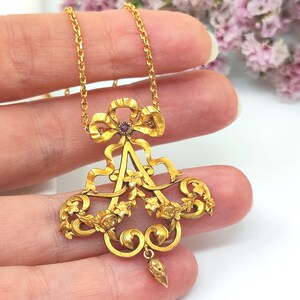 May include: A gold-colored pendant necklace with an ornate design. The pendant features a bow, floral accents, and a central letter-like shape, with a small red gemstone. The necklace chain is also gold.