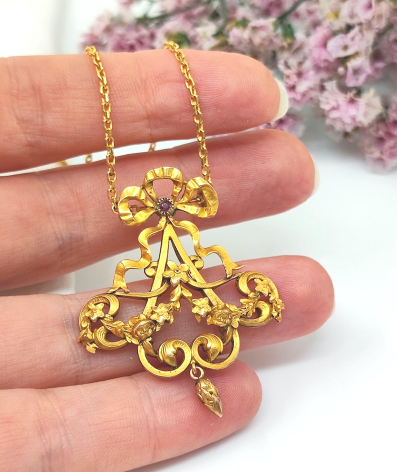 May include: A gold-colored pendant necklace with an ornate design. The pendant features a bow, floral accents, and a central letter-like shape, with a small red gemstone. The necklace chain is also gold.