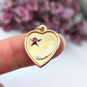May include: A gold heart-shaped pendant with a star and a small red gemstone. The pendant has a textured surface with engraved details and a loop for a chain. The word "amour" is engraved on the pendant.