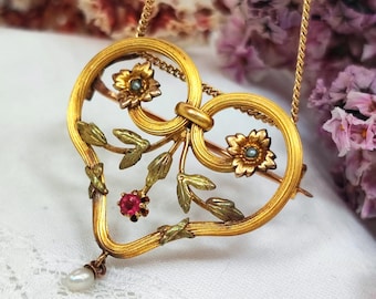 Peut inclure: Pendentif doré en forme de cœur avec des motifs floraux et de feuilles. Le pendentif comporte deux fleurs avec de petites pierres bleu-vert, une pierre rouge et une perle pendante. La chaîne est également dorée.