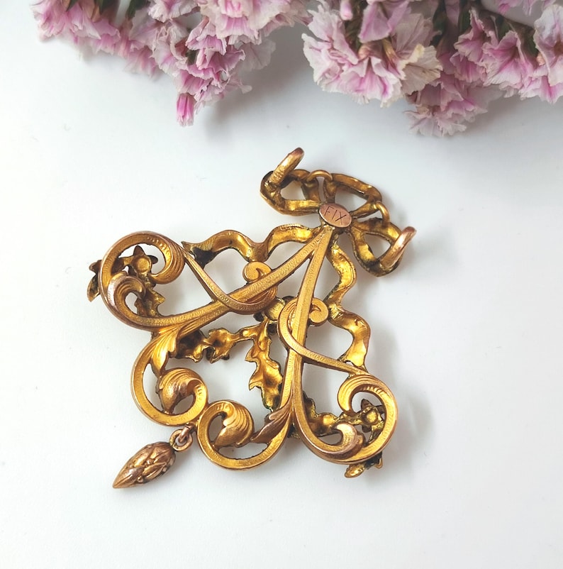 May include: Ornate gold-toned brooch featuring the letter "A" in a decorative script, intertwined with floral and ribbon elements. A small, dangling pendant hangs from the bottom. The brooch is set against a white background, with pink flowers in the upper corner.