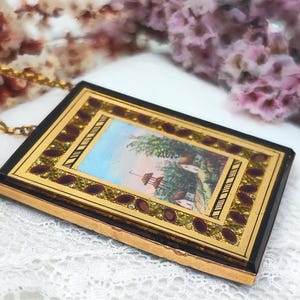 May include: A rectangular pendant with a gold chain. The pendant has a black border, a gold frame, and a miniature painting of a landscape with a house and trees. The frame is decorated with red and gold accents.
