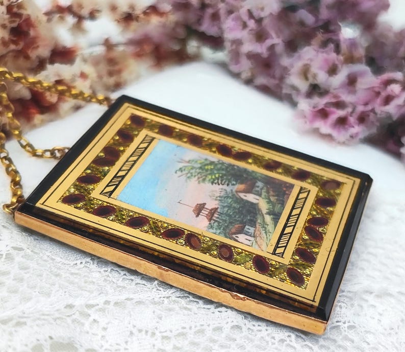 May include: A rectangular pendant with a gold chain. The pendant has a black border, a gold frame, and a miniature painting of a landscape with a house and trees. The frame is decorated with red and gold accents.
