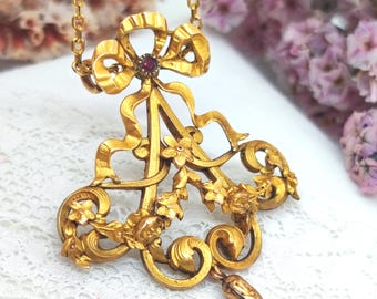 Peut inclure: Collier pendentif doré avec un nœud orné d'une pierre rouge au centre. Le pendentif présente des détails floraux et des rubans, ainsi qu'une chaîne délicate. Le collier est probablement vintage ou antique.