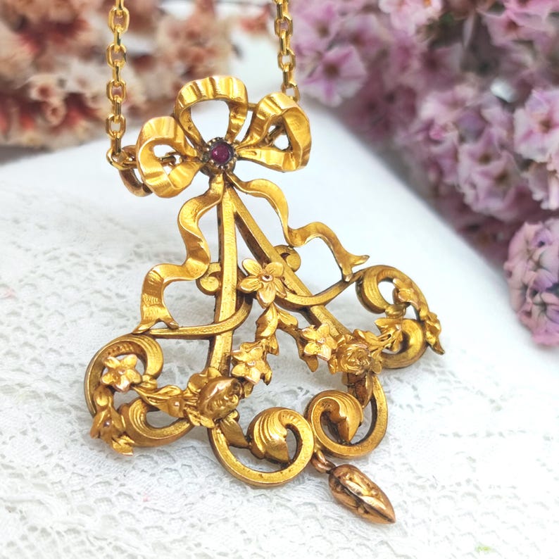 May include: Gold-toned pendant necklace featuring a bow design with a central red gemstone. The pendant has floral and ribbon details, and a delicate chain. The necklace is likely vintage or antique.