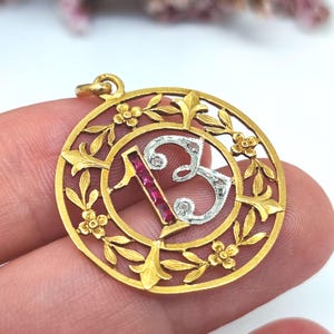 May include: A circular gold pendant featuring an openwork floral frame surrounding the number 13. The number one is inlaid with rectangular red stones, while the number three is crafted from silver-toned metal with small embedded clear stones.