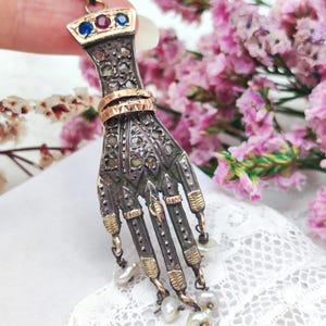 Op de afbeelding: Een vintage hanger in de vorm van een hand, gemaakt van zilver en goud. De hand heeft ingewikkelde details, kleine edelstenen en bungelende parels. De hanger heeft een gouden beugel met blauwe en rode edelstenen.