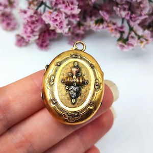 May include: Oval gold locket with ornate detailing. The locket features a central design with a cross-like structure and a detailed face. The locket has a loop at the top for a chain. The background includes pink flowers.