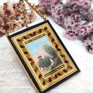 May include: A rectangular, antique miniature painting with a gold frame and a black border. The painting depicts a landscape scene with a blue sky, trees, and small buildings. The frame is adorned with red and gold accents, and it hangs from a gold chain.