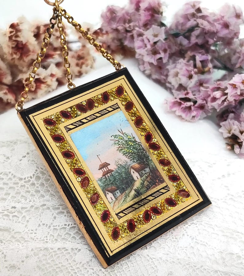 May include: A rectangular, antique miniature painting with a gold frame and a black border. The painting depicts a landscape scene with a blue sky, trees, and small buildings. The frame is adorned with red and gold accents, and it hangs from a gold chain.