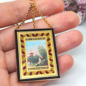 May include: A rectangular pendant with a miniature landscape painting. The pendant has a black frame with gold accents and red gemstone embellishments. It hangs from a gold chain.
