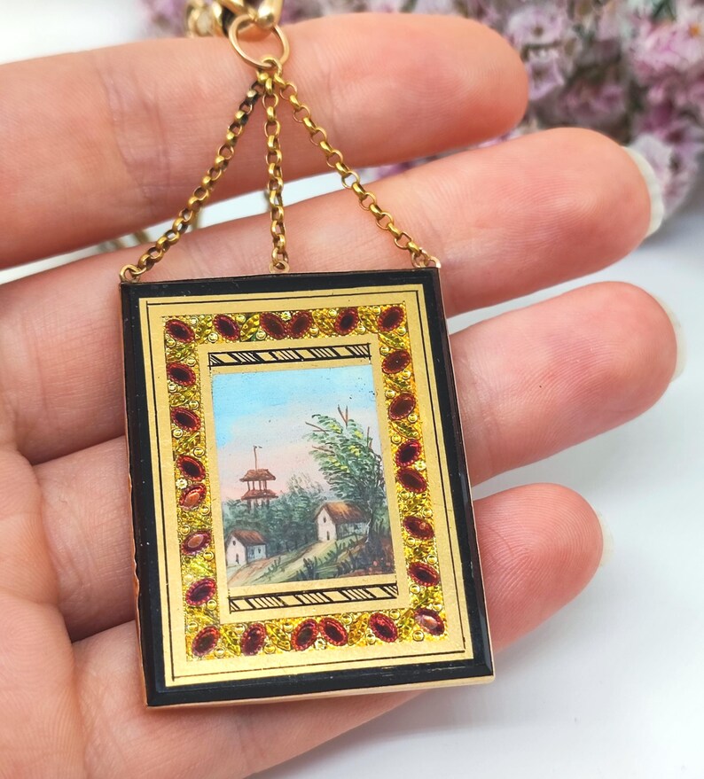 May include: A rectangular pendant with a miniature landscape painting. The pendant has a black frame with gold accents and red gemstone embellishments. It hangs from a gold chain.