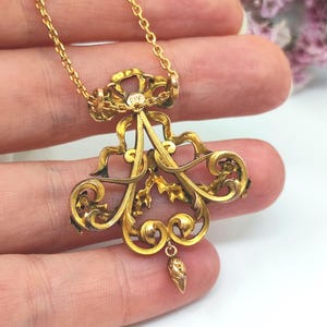 May include: A gold-colored pendant necklace with an ornate, scrollwork design. The pendant features a central floral motif with a small, dangling teardrop-shaped element. The chain is delicate and gold, and the pendant has the text "FIX".