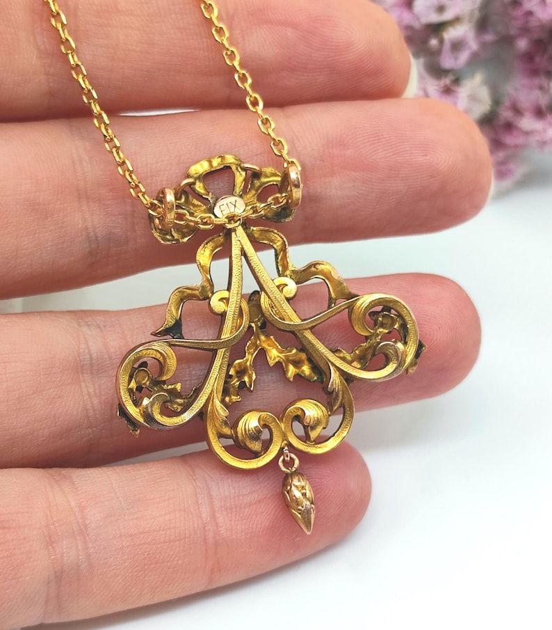 May include: A gold-colored pendant necklace with an ornate, scrollwork design. The pendant features a central floral motif with a small, dangling teardrop-shaped element. The chain is delicate and gold, and the pendant has the text "FIX".