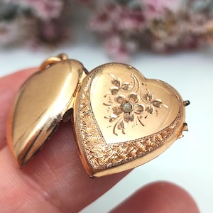 Op de afbeelding: Een goudkleurige, hartvormige medaillonhanger met een gegraveerd bloemmotief op de voorzijde. Het medaillon staat op een kier en toont een glad, effen binnenoppervlak aan de linkerzijde en een textuurrijke, gedetailleerde rand aan de rechterkant.