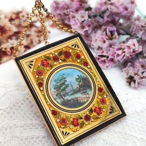 May include: A rectangular, gold-toned pendant with a black border and a small, round painting of a landscape. The pendant is decorated with red roses and small, sparkling accents. A delicate gold chain suspends the pendant.