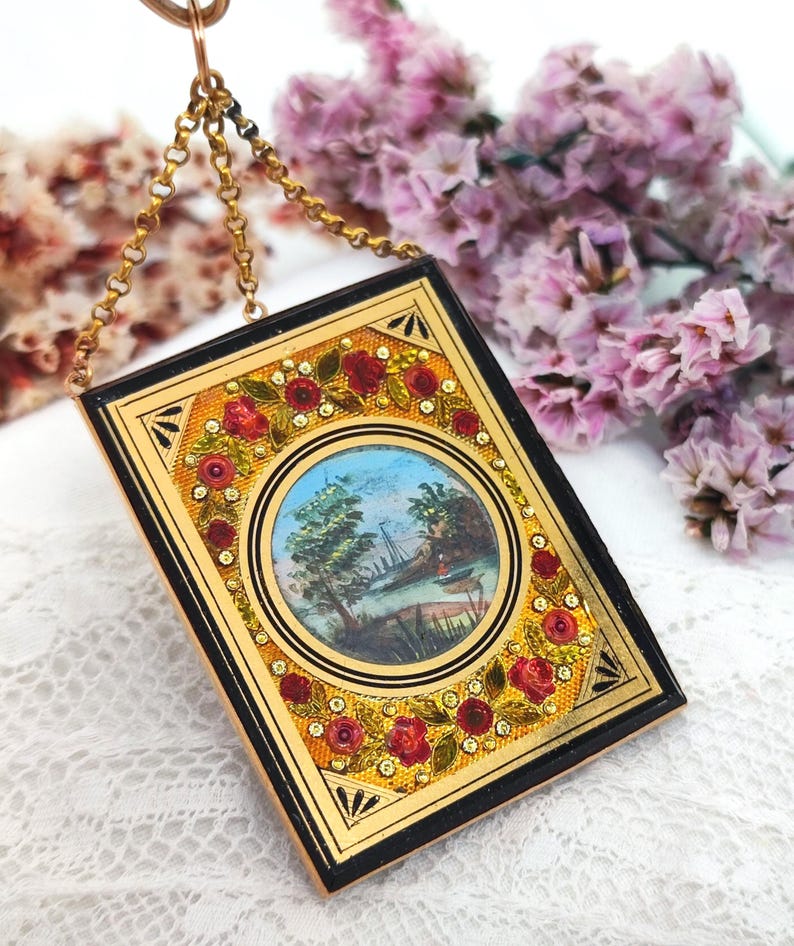 May include: A rectangular, gold-toned pendant with a black border and a small, round painting of a landscape. The pendant is decorated with red roses and small, sparkling accents. A delicate gold chain suspends the pendant.