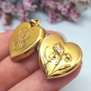 Puede incluir: Dos relicarios de oro en forma de corazón. Un relicario presenta un monograma grabado intrincado con letras en cursiva. El otro relicario muestra un relieve detallado de una flor de iris con hojas y un tallo sobre su superficie lisa.