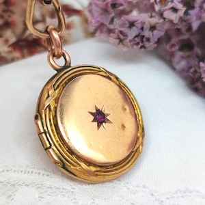 Puede incluir: Un relicario ovalado de color dorado con una gema en forma de estrella de color rubí en el centro. El relicario está suspendido de una cadena a juego. El relicario tiene un diseño ornamentado en los bordes.