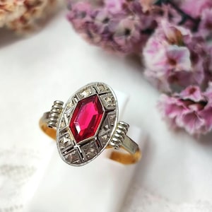 Peut inclure: Bague de style vintage avec une grosse pierre rouge rubis facettée dans un sertissage ovale. La bague présente un motif filigrané argenté autour du rubis et de petites pierres claires. La monture est dorée.