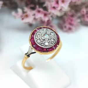 May include: A gold ring featuring a circular head with a central cluster of small round diamonds set in white metal, surrounded by a halo of square-cut vibrant red rubies. The band is thin and gold, connecting to the setting with a metal accent.