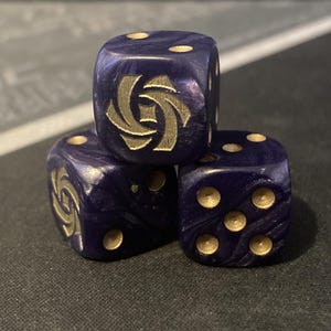 May include: Three dark blue dice with gold-colored pips and a gold emblem on one face. The dice are stacked, with the top die displaying the emblem. The dice have a marbled appearance.