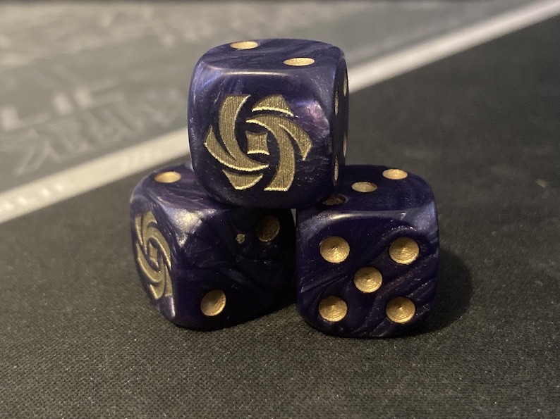 May include: Three dark blue dice with gold-colored pips and a gold emblem on one face. The dice are stacked, with the top die displaying the emblem. The dice have a marbled appearance.