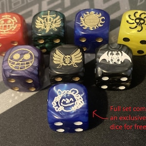 Op de afbeelding: Een set van zeven kleurrijke dobbelstenen met verschillende gouden symbolen. De dobbelstenen zijn rood, groen, blauw, geel, paars en zwart. De tekst op de afbeelding luidt: "Complete set wordt geleverd met een exclusieve Kuma dobbelsteen gratis!"