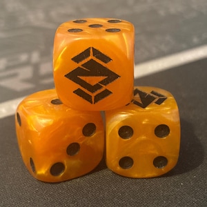 May include: Three orange dice with black pips and a black geometric design on one face. The dice have a marbled appearance and are arranged in a small pyramid shape. The dice are for a board game.