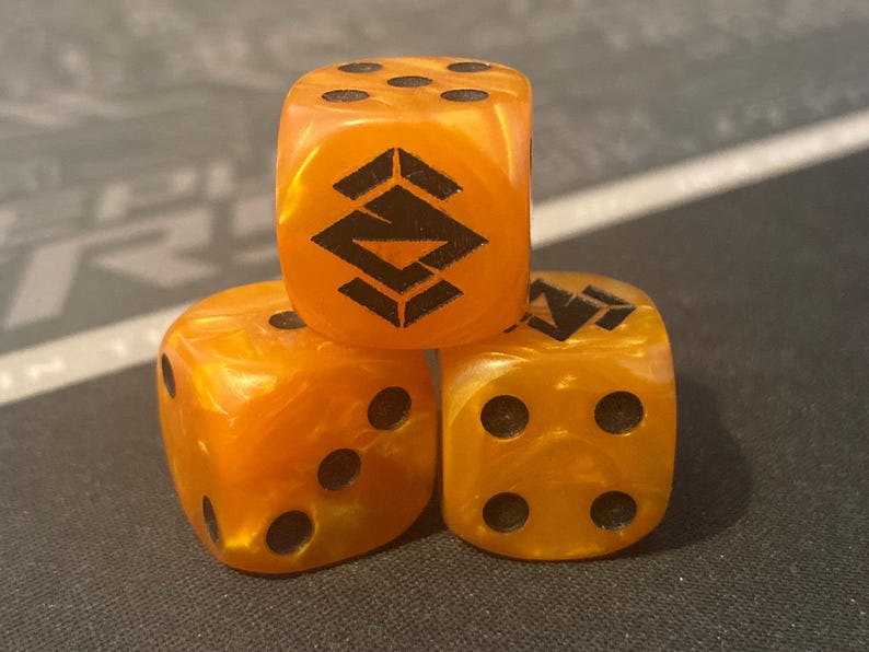 May include: Three orange dice with black pips and a black geometric design on one face. The dice have a marbled appearance and are arranged in a small pyramid shape. The dice are for a board game.
