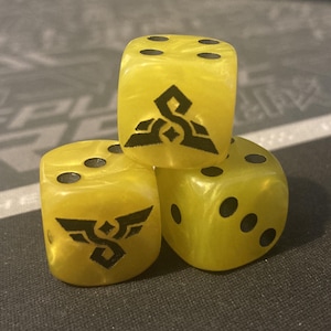 May include: Three yellow dice with black pips and black graphic designs. The dice are stacked on top of each other. The dice have a marbled appearance.