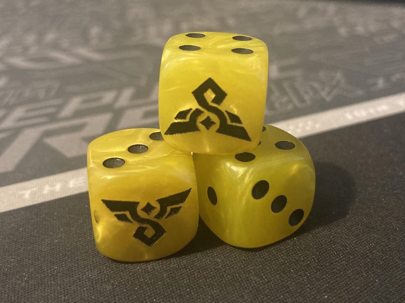 May include: Three yellow dice with black pips and black graphic designs. The dice are stacked on top of each other. The dice have a marbled appearance.