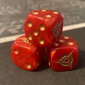 May include: Three red dice with gold-colored pips and a gold emblem on each face. The dice are stacked on top of each other, creating a visually interesting arrangement. The dice are made of a translucent material.