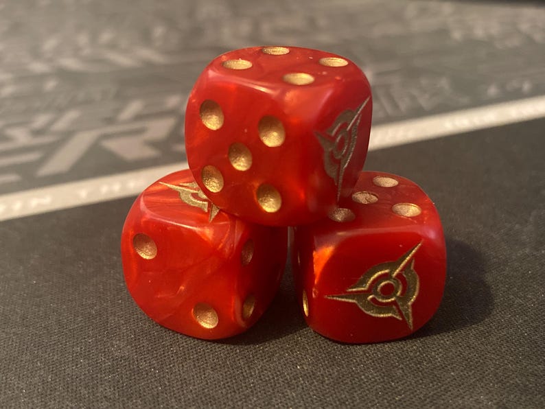 May include: Three red dice with gold-colored pips and a gold emblem on each face. The dice are stacked on top of each other, creating a visually interesting arrangement. The dice are made of a translucent material.