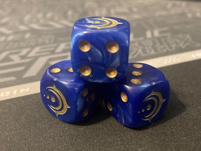 May include: Three blue dice with gold markings and symbols. The dice are stacked, showing different numbers of dots and a crescent moon design. The dice have a marbled effect and are set against a dark background.