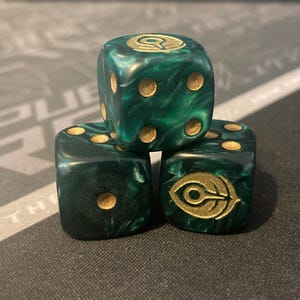 May include: Three green and gold dice stacked on top of each other. The dice have gold pips and a gold symbol on one face. The dice are a marbled green color.