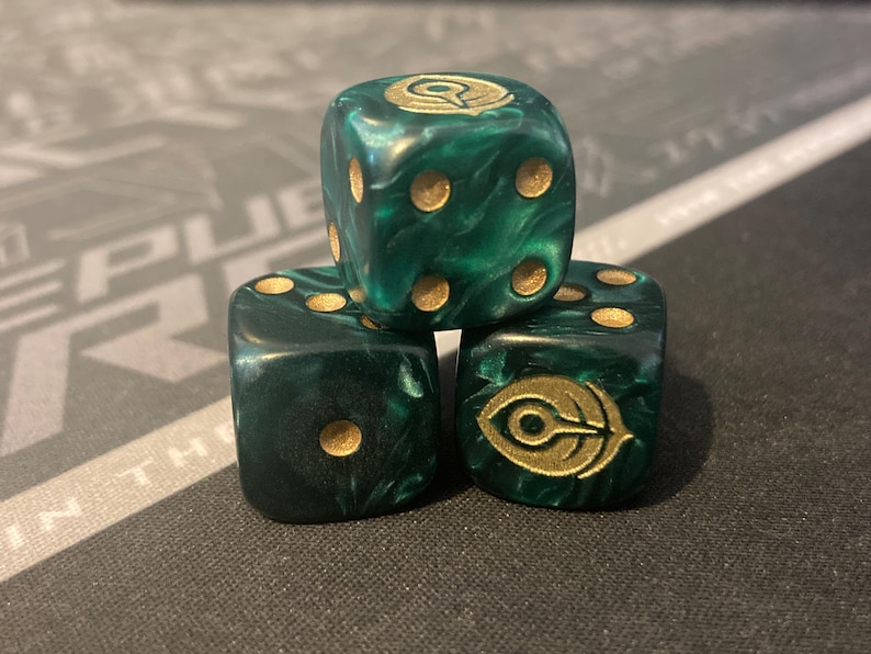 May include: Three green and gold dice stacked on top of each other. The dice have gold pips and a gold symbol on one face. The dice are a marbled green color.