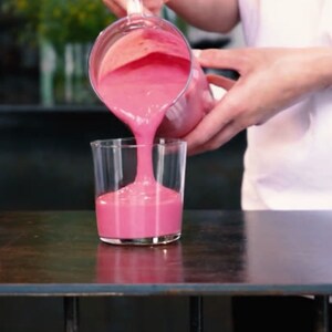 Puede incluir: Un líquido rosa brillante se vierte de una jarra transparente a un vaso transparente. La escena se desarrolla sobre una superficie de madera oscura, con un fondo borroso. El líquido se vierte en el vaso.