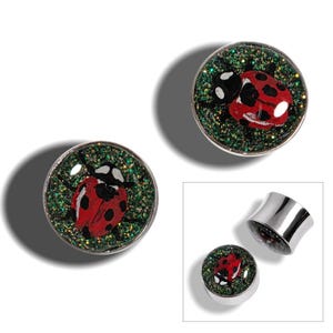 May include: A pair of silver-colored ear gauges with a ladybug design. The ladybug is red with black spots, set against a green glitter background. One gauge is shown separately, displaying its hollow interior.