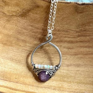 May include: Silver pendant necklace featuring a wire-wrapped design. The pendant has a teardrop shape with a large, faceted purple gemstone at the bottom and small white gemstones above. The necklace chain is also silver.
